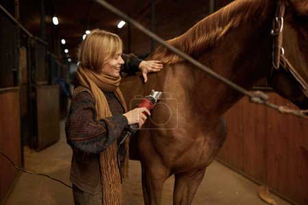 Foto de Joven mujer satisfecha aseo caballo usando afeitadora eléctrica en establo rancho. Concepto de cría veterinaria, equina y cuidado de los animales domésticos - Imagen libre de derechos