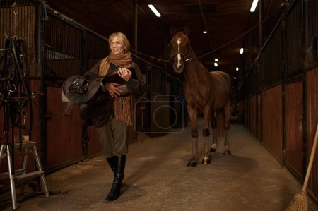 Foto de Female horse rider walking with harness in stable. Young adult woman rancher going with saddle in hands and smiling - Imagen libre de derechos