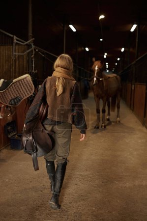 Foto de View from back on walking horsewoman rancher carrying saddle in hand. Preparation for training with thoroughbred horse. Stable interior - Imagen libre de derechos