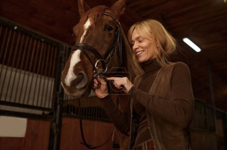 Foto de Happy smiling woman rider putting bridle horsey muzzle while standing in stable. Harnessing stallion before riding - Imagen libre de derechos