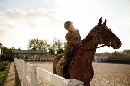 Foto de Attractive smiling female horse rider training in outdoor paddock. Riding club and equine caring concept - Imagen libre de derechos
