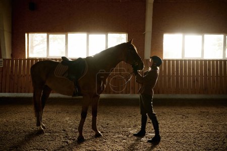 Foto de Mujer jinete caballo enganchado en establo paddock interior. Joven semental de formación en el club de equitación - Imagen libre de derechos
