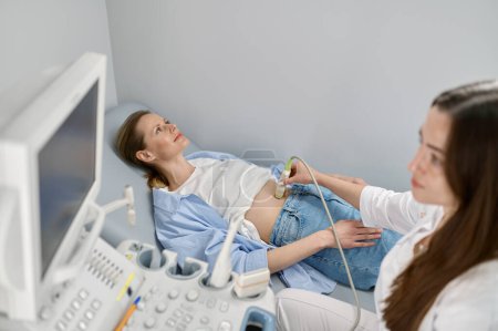 Foto de Médico profesional examinando a su paciente haciendo un ultrasonido abdominal utilizando un dispositivo de escáner de ecografía. Concepto de confirmación temprana del embarazo - Imagen libre de derechos