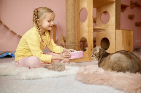 Téléchargez les photos : Petite fille mignonne offrant au chat de la nourriture ou de l'eau dans une assiette en plastique tout en passant du temps à l'abri des animaux - en image libre de droit