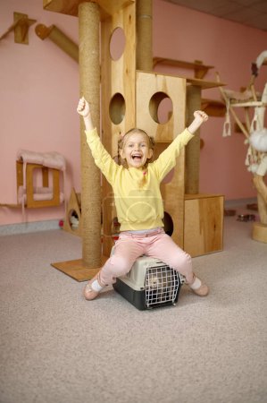 Téléchargez les photos : Petite fille joyeuse assise sur une cage avec chat au-dessus d'une aire de jeux abritée. Enfant appréciant compagnie d'avoir animal de compagnie et d'apprendre la responsabilité des soins des animaux - en image libre de droit