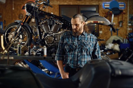 Photo for Portrait of bearded brutal biker at motorcycle in garage workshop. Motorbike service - Royalty Free Image