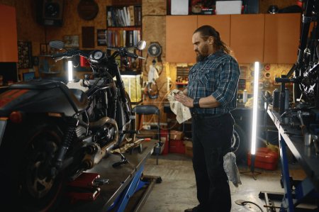 Téléchargez les photos : Brutal motard essuyant ses mains tout en réparant le moteur de moto à l'atelier de réparation. Mécanicien masculin barbu travaillant sur le concept d'entretien de moto - en image libre de droit
