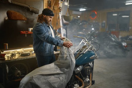 Photo for Mature male biker covering his new motorbike with blanket after repair work in garage workshop - Royalty Free Image