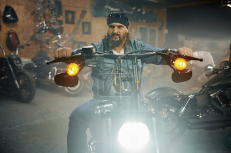 Foto de Hombre barbudo guapo probando moto nueva en la tienda de garaje. Motociclista elegir nuevo vehículo en el taller de automóviles - Imagen libre de derechos