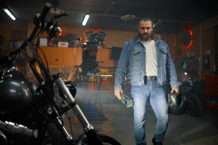 Téléchargez les photos : Portrait de motard barbu brutal regardant la moto dans un atelier de garage. Service de moto - en image libre de droit