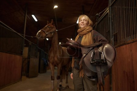 Foto de Joven jinete adulto sonriente de pie y con silla de montar en la mano sobre su caballo en el establo. Antes de montar deporte - Imagen libre de derechos