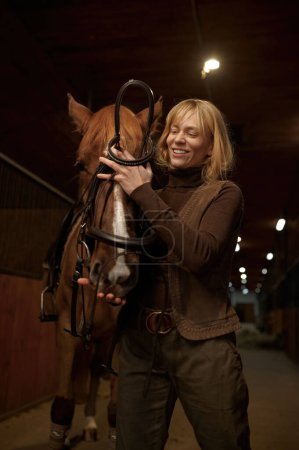 Foto de Mujer jinete aprovechando caballo en establo de club de equitación o rancho. hembra jinete poniendo arnés en semental cara vista frontal disparo - Imagen libre de derechos
