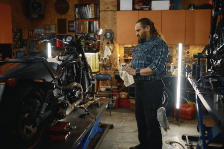 Foto de Brutal motociclista limpiándose las manos mientras reparaba el motor de la motocicleta en el taller de reparación. Mecánico barbudo trabajando en el concepto de mantenimiento de motocicletas - Imagen libre de derechos