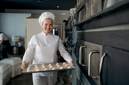 Foto de Retrato de mujer panadera en bandeja de transporte uniforme con galletas en forma de masa y preparada para hornear. Producción pastelera - Imagen libre de derechos