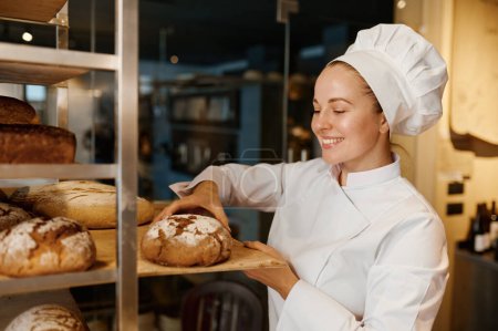 Foto de Hermosa mujer panadero en uniforme de control de pan recién horneado en bandeja en la cocina de panadería moderna - Imagen libre de derechos