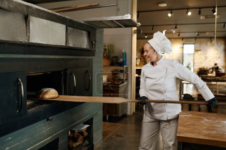 Foto de Trabajadora de panadería joven usando pala de madera para tomar pan fresco del horno de cocina caliente. Producción industrial de productos pasteleros - Imagen libre de derechos