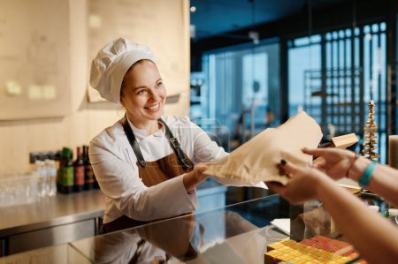 Photo for Bakery worker selling fresh tasty pastry and bread in bakery shop. Focus on young woman giving eco paper pack with order to customer - Royalty Free Image