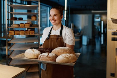 Photo for Pretty woman baker holding variety of baked bread on tray standing over bakery house background - Royalty Free Image