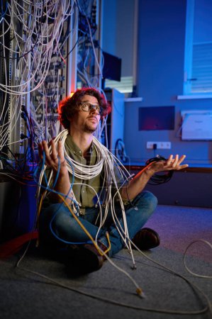 Foto de Técnico triste y perplejo que sostiene muchos cables en la mano mirando hacia arriba tratando de resolver el problema con la conexión a Internet en la sala de servidores del centro de datos - Imagen libre de derechos