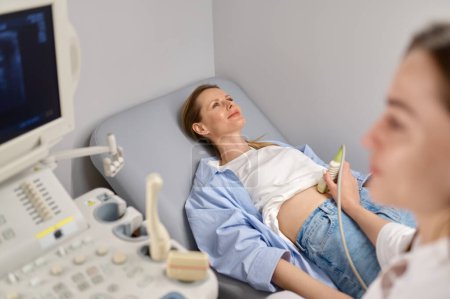 Téléchargez les photos : Médecin professionnel examinant son patient faisant une échographie abdominale à l'aide d'un appareil de scanner d'échographie. Concept de confirmation précoce de grossesse - en image libre de droit