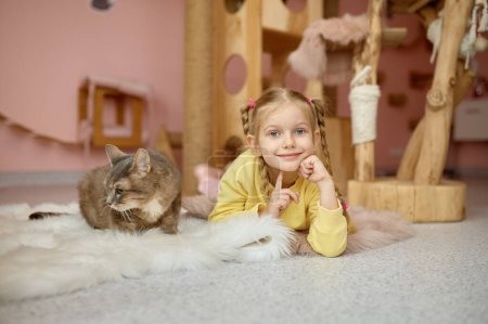 Téléchargez les photos : Portrait de fille gaie couchée sur le sol avec chat moelleux sur fond d'abri pour animaux. Enfant et chaton - en image libre de droit