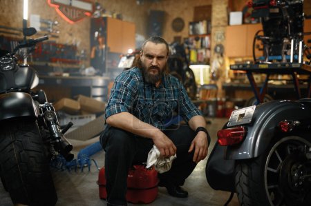 Téléchargez les photos : Homme mûr barbu motard nettoyage moto dans le poste de travail de garage. Portrait de mécanicien assis près de la moto - en image libre de droit