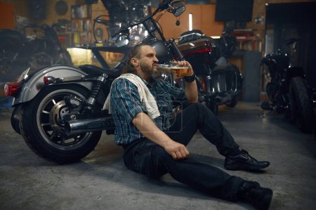 Foto de Hombre cansado motociclista beber cerveza de botella después de un duro trabajo de reparación en el garaje. Retrato del hombre maduro sentado en su motocicleta - Imagen libre de derechos