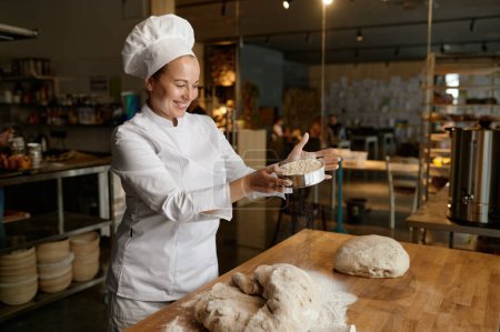 Foto de Feliz mujer sonriente panadero en uniforme siembra harina a través del tamiz de cocina en la masa. Cocina de repostería en el trabajo - Imagen libre de derechos