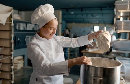 Photo for Professional female confectioner wearing white uniform putting flour into big metal bowl standing near table in confectionery - Royalty Free Image