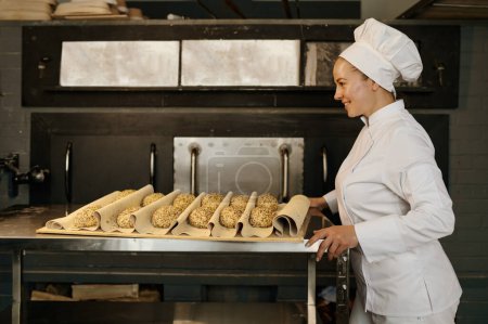 Téléchargez les photos : Jeune femme boulanger tirant un plateau avec des petits pains crus avec des graines de sésame préparées pour la cuisson vue de côté - en image libre de droit