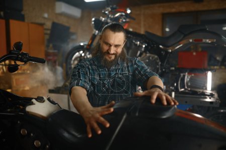 Foto de Motociclista satisfecho mirando a su moto sensación de emoción y la mano en el nuevo asiento de cuero. Garaje taller interior - Imagen libre de derechos