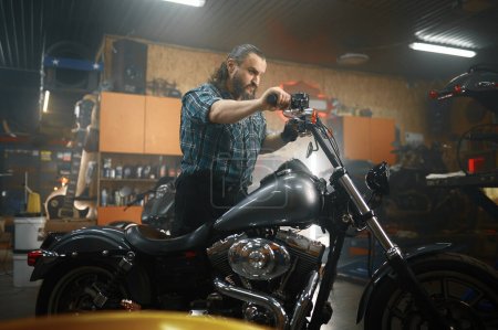 Téléchargez les photos : Homme mûr motard essayant sa nouvelle moto dans le poste de travail du garage. Homme adulte mécanicien prêt pour la course - en image libre de droit