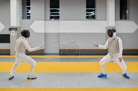 Photo for Two swordsmen wearing uniform and protective helmet fighting duel during training in sport gym room. Fencing club - Royalty Free Image