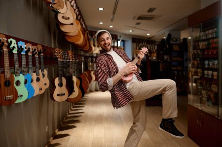 Photo for Overjoyed funny guitarist playing ukulele guitar posing for camera standing at musical instrument shop store - Royalty Free Image
