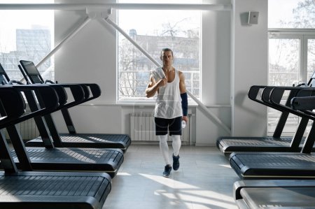 Téléchargez les photos : Bodybuilder mâle musculaire ayant pause après cardio. Homme confiant athlète sur tapis roulants situé dans l'intérieur de la salle de gym - en image libre de droit