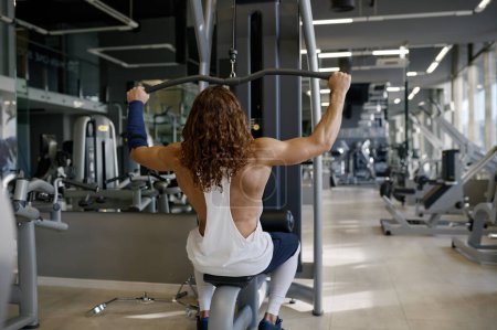 Foto de Hombre atlético joven motivado que bombea los músculos que tienen entrenamiento en la máquina de ejercicio del gimnasio que levanta diversos pesos. Vista trasera - Imagen libre de derechos