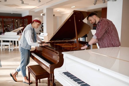 Foto de Joven pareja de músicos eligiendo un nuevo instrumento para la banda familiar. Hipster pianista hombre y mujer examinando el piano en la tienda de música moderna - Imagen libre de derechos