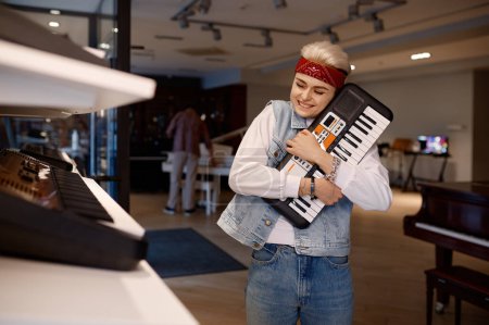 Foto de Cliente femenino feliz sosteniendo un nuevo teclado de sintetizador eléctrico sintiendo satisfacción y placer. Mujer alegre músico disfrutar de compras en la tienda de música - Imagen libre de derechos