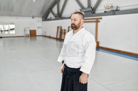 Foto de Entrenador serio aikido usando kimono posando sobre fondo de gimnasio deportivo vacío. Concepto de artes marciales y formación - Imagen libre de derechos