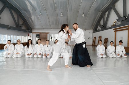Foto de Instructor de Aikido enseñando luchadores adolescentes durante el entrenamiento en grupo en el gimnasio de la escuela de deporte. Maestra y chica de artes marciales en kimono mostrando técnica de protección para estudiantes - Imagen libre de derechos