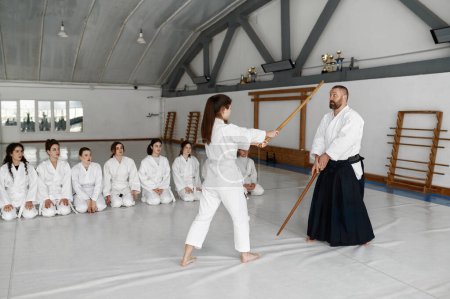 Foto de Chica y maestro aikido luchando con espada de madera bokken durante la clase de artes marciales. Adolescente grupo femenino sentado cerca en el suelo y mirando - Imagen libre de derechos