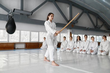 Foto de Chica joven luchando con espada de madera en el entrenamiento aikido en la escuela de artes marciales. Adolescente luchadora en kimono blanco mostrando técnica a sus compañeros de clase - Imagen libre de derechos