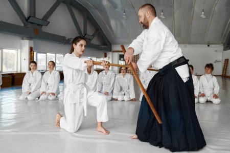 Téléchargez les photos : Sensei avec une jeune étudiante combattant avec une épée en bois démontrant des compétences d'aikido à un groupe de combattantes adolescentes - en image libre de droit