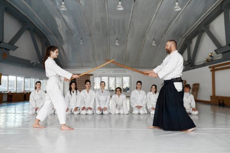 Photo for Martial arts master teaching young student to fight with wooden sword. Group of female teenagers watching battle and learning - Royalty Free Image