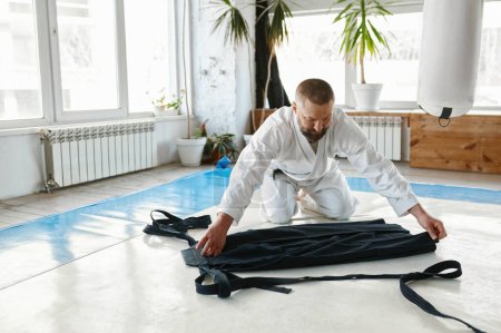 Téléchargez les photos : Maître aikido mature pliant kimono hakama après la classe d'entraînement aux arts martiaux à la salle de gym de l'école sportive professionnelle - en image libre de droit