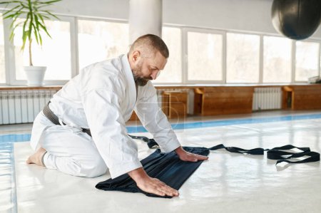 Téléchargez les photos : Maître aikido mature pliant kimono hakama après la classe d'entraînement aux arts martiaux à la salle de gym de l'école sportive professionnelle - en image libre de droit