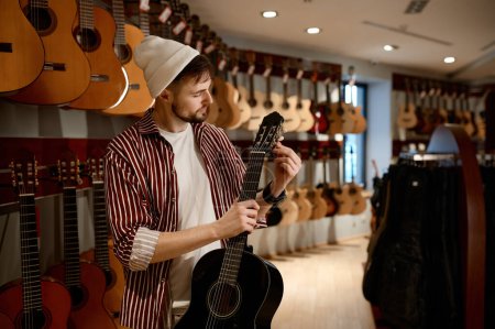 Téléchargez les photos : Jeune client masculin choisissant la guitare acoustique à la boutique. Hipster gars toucher le cou de l'instrument debout à proximité rack d'équipement - en image libre de droit