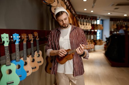 Foto de Joven músico hipster hombre comprar guitarra ukelele en tienda de instrumentos musicales. Hobby, concepto de negocios y comercio - Imagen libre de derechos