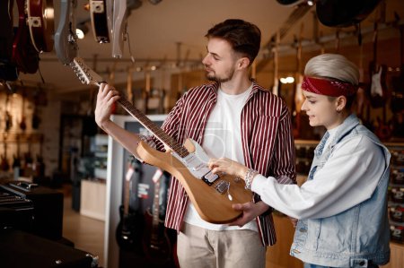 Téléchargez les photos : Jeune couple hipster à la recherche d'une nouvelle guitare électrique pour la performance en choisissant un instrument à cordes dans un magasin d'art professionnel - en image libre de droit