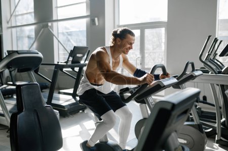 Photo for Healthy smiling sportsman using elliptical machine at gym fitness center. Happy motivated male bodybuilder doing aerobic exercise for slim and firm healthy body - Royalty Free Image